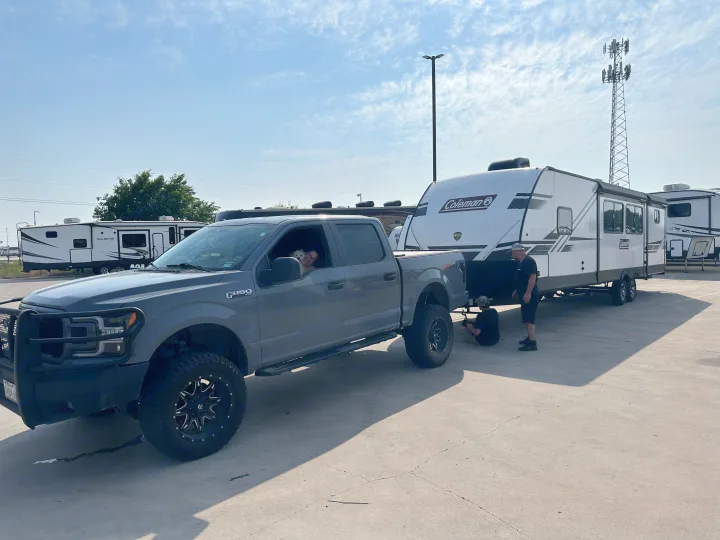 RV rental setup: A gray Ford F-150 truck hitches to a Coleman travel trailer. Two men work on the hitch; a person and dog watch from the truck.. Dutchmen Coleman Light 2023