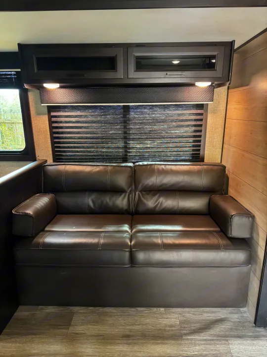 Comfortable RV rental living area: dark faux leather sofa with white stitching, window with blinds, lighted overhead cabinets, wood-look floor.. Jayco Jay Flight 2022