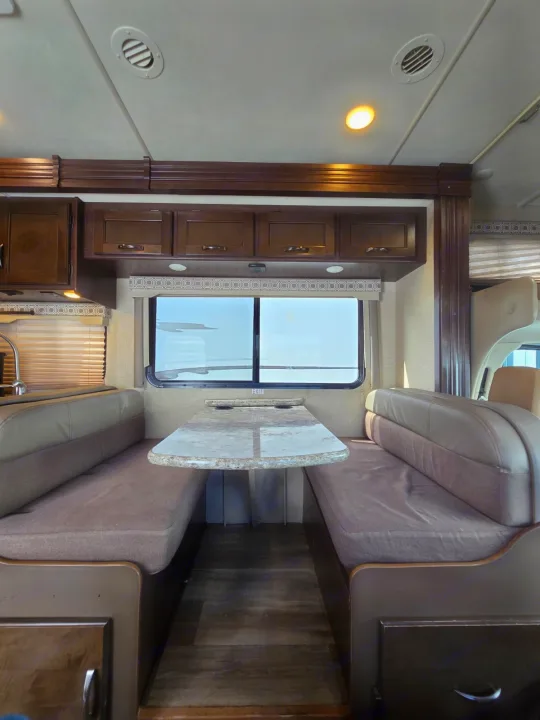 This RV rental interior showcases a comfy dinette with upholstered benches, a table, overhead cabinets, and a window view.. Thor Four Winds 2019