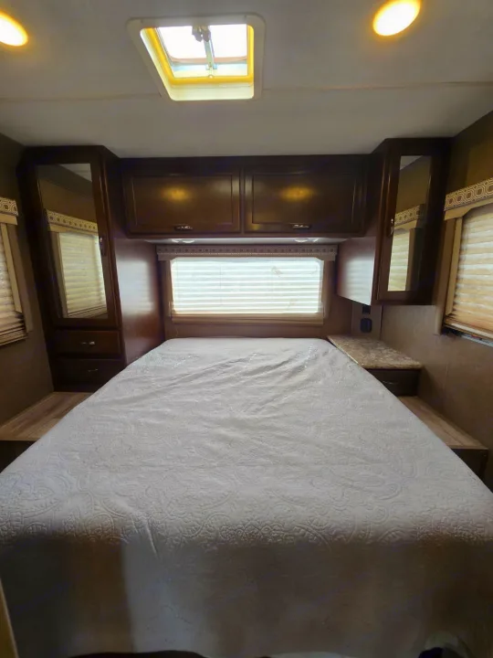 RV rental bedroom: a comfy bed with quilted spread, overhead cabinets, side mirrors, window, and a ceiling skylight.. Thor Four Winds 2019