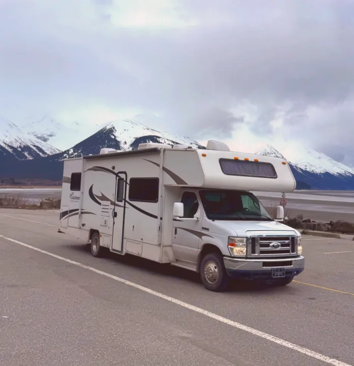 A white Class C rv rental is parked roadside, with majestic snow-capped mountains & a calm bay under a cloudy sky.. Coachmen Freelander 2014