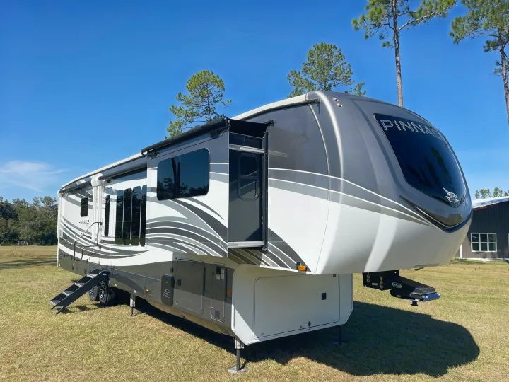 Pristine gray and white Pinnacle fifth-wheel RV rental with slide-outs extended, nestled in a sunny, grassy setting. Ideal for travel.. Jayco Pinnacle 2023