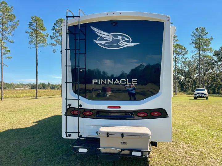 Rear of a white RV rental, showing a ladder, "Pinnacle by Jayco" logo, and a Yeti cooler on a carrier, parked in a sunny, grassy field.. Jayco Pinnacle 2023