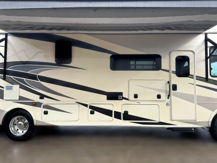 Side view of a beige and grey RV rental with an extended awning, multiple windows, and storage compartments along its side.. Coachmen Encore 2021