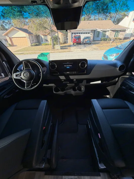 Inside a Mercedes-Benz RV rental: the front cabin showing dashboard, steering wheel, and black leather seats, facing a residential street.. Cahaba Cahaba Class B 2022