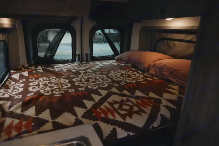 RV rental interior: A cozy bed with a patterned blanket, pink pillows, and windows, perfect for mobile sleeping.. Winnebago Winnebago 2026