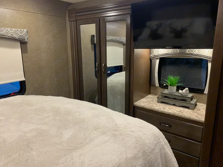 Cozy RV rental bedroom: soft white comforter, mirrored closet, TV, and decorative items on a counter.. Thor Chateau 2020