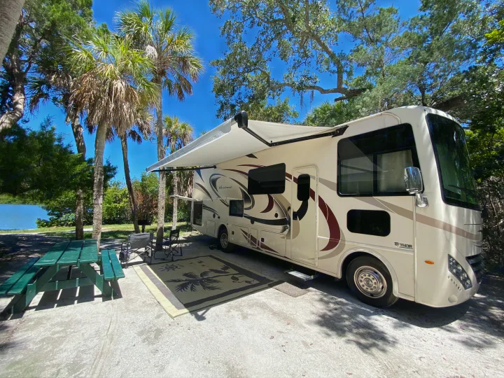 RV rental parked at a scenic lakeside campsite with palm trees, a picnic table, and an extended awning.. Thor Hurricane 2018