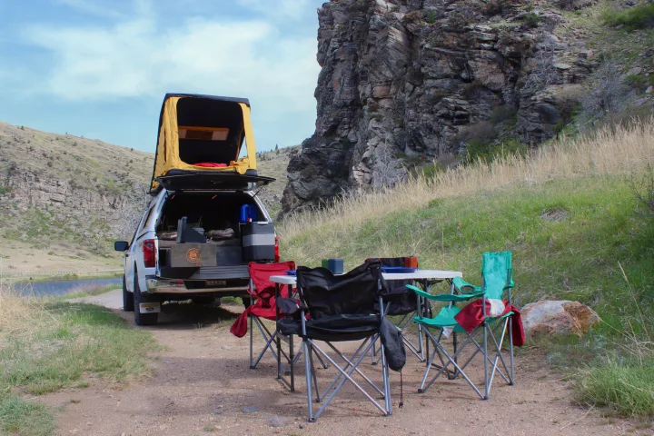 A truck with a rooftop tent, camping chairs, and table set up by a scenic lake. An ideal outdoor adventure for any RV rental!. Ford Ford F150 2024
