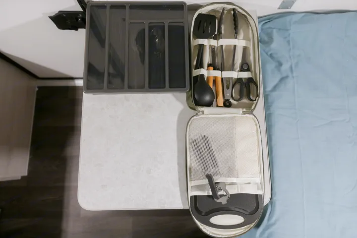 RV rental kitchen: An open case displays spatula, ladle, tongs, skewers, scissors, and cutting board next to a cutlery organizer.. A Liner Scout 2026