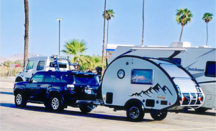 Dark SUV towing a compact teardrop RV. A larger Dutchmen RV is parked nearby, ideal for an RV rental trip.. nuCamp T@B 2022