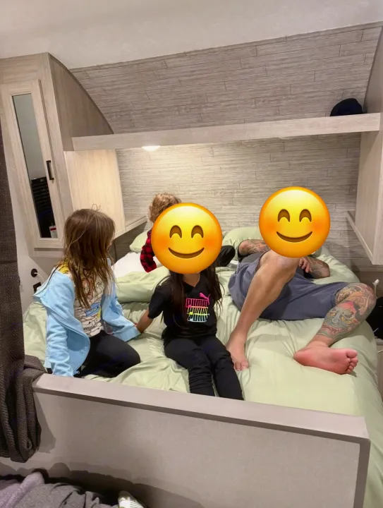 Family relaxes inside an RV rental. Kids and an adult with tattoos lounge on a large bed in the modern, wood-paneled interior.. Coachmen Catalina Sumit 8 2025