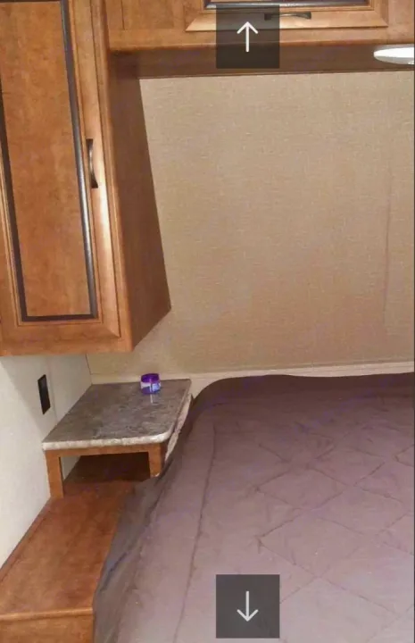 RV rental bedroom with a cozy bed, small nightstand holding a purple container, and wooden overhead cabinets.. Crossroads RV Zinger 2017