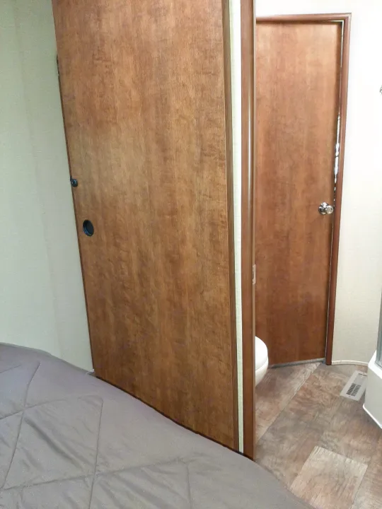 Inside an rv rental, a bedroom area with a gray bed leads to a compact bathroom through a partially open wooden door, revealing a toilet.. Crossroads RV Zinger 2017