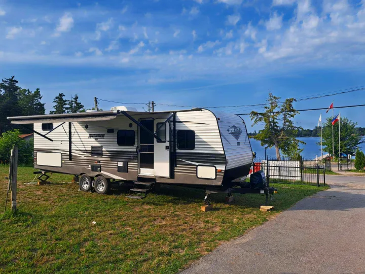 Bright travel trailer RV rental set up on a grassy site by a serene lake under a blue sky, perfect for a relaxing getaway.. Avenger ATI 2019