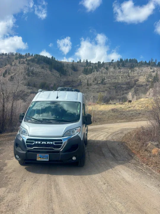 White Ram RV rental van on a dirt road in Wyoming, with mountains and a picnic area under a blue sky.. Thor Scope 2024