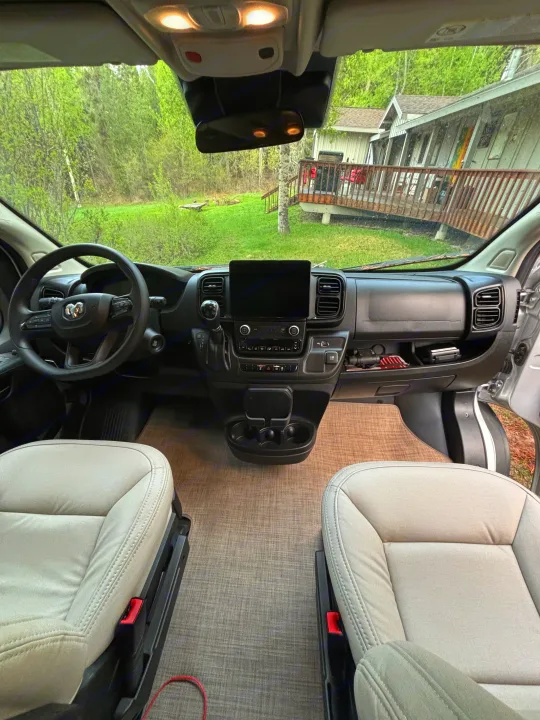 Inside this RV rental: front cabin with comfortable seats, a modern dashboard, and a scenic forest view outside a home.. Thor Scope 2024