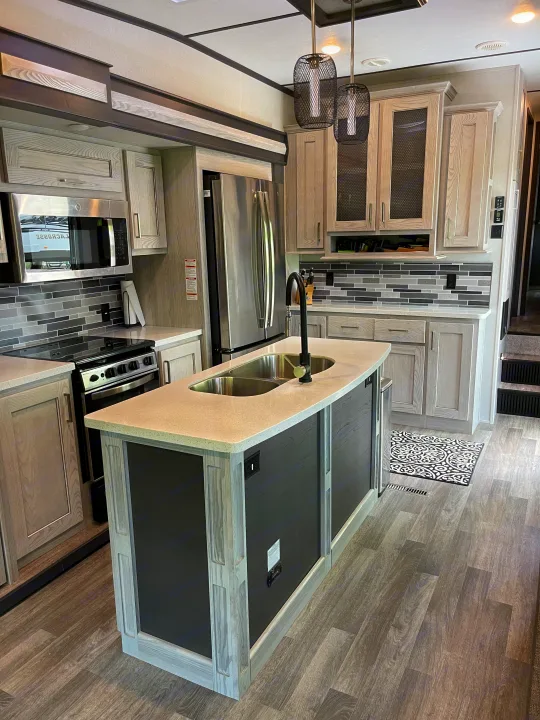 Modern RV rental kitchen: light wood-grain cabinets, island with double sink, black faucet, stainless appliances, and gray flooring.. Keystone RV Montana High Country 2021