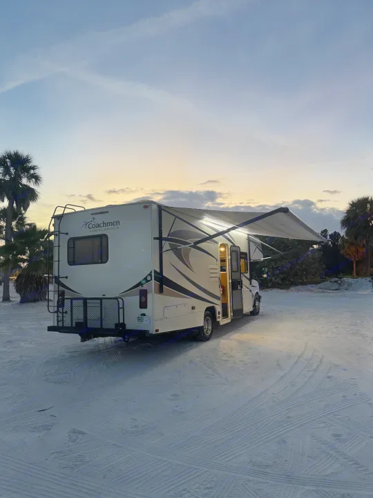 Serene twilight camping with an RV rental parked on soft sand, its awning extended & lights glowing. Palm trees frame the colorful sky.. Coachmen Freelander 26 RS 2017