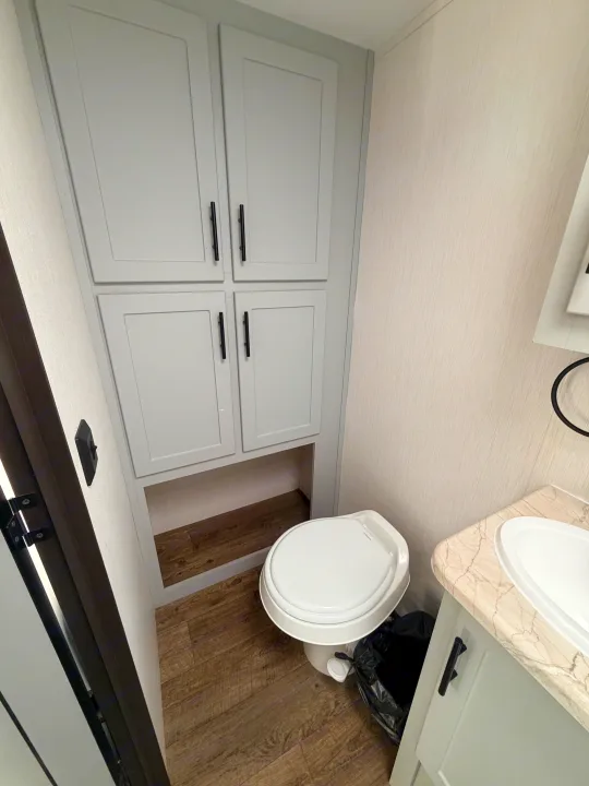 Inside an RV rental bathroom, featuring light gray cabinets above a white toilet, sink vanity, and wood-look floor.. Forest River Vibe 2021
