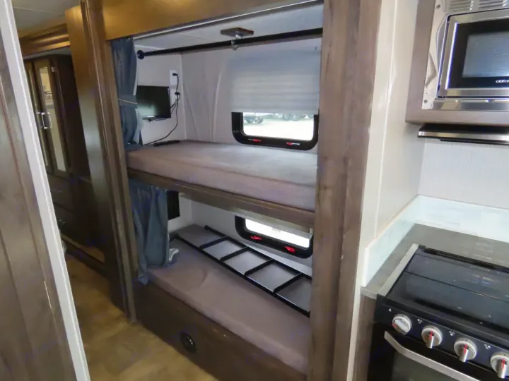 RV rental interior features modern wood-grain bunk beds with privacy curtains, small windows, a mounted TV, and a compact kitchen with stove/microwave.. Coachmen PURSUIT 31BH 2023