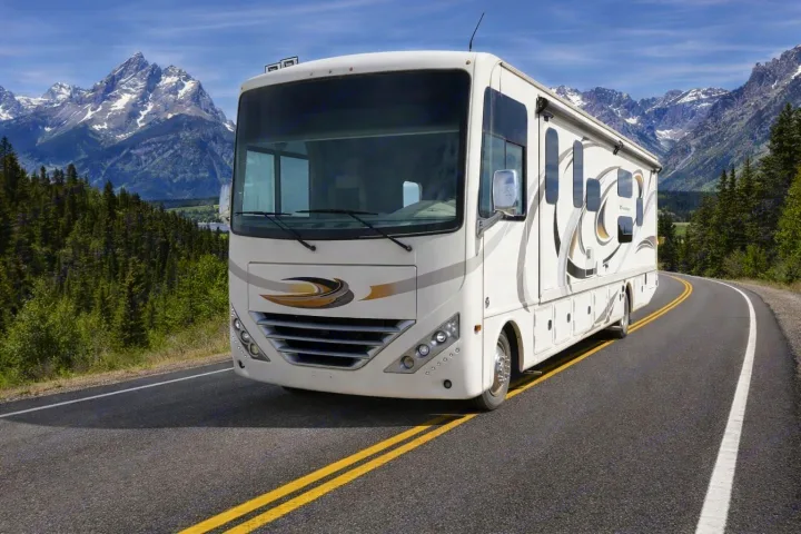 Experience epic travels with an RV rental! A large white RV drives on a scenic mountain road, surrounded by forests and snow-capped peaks.. Thor Hurricane 2017