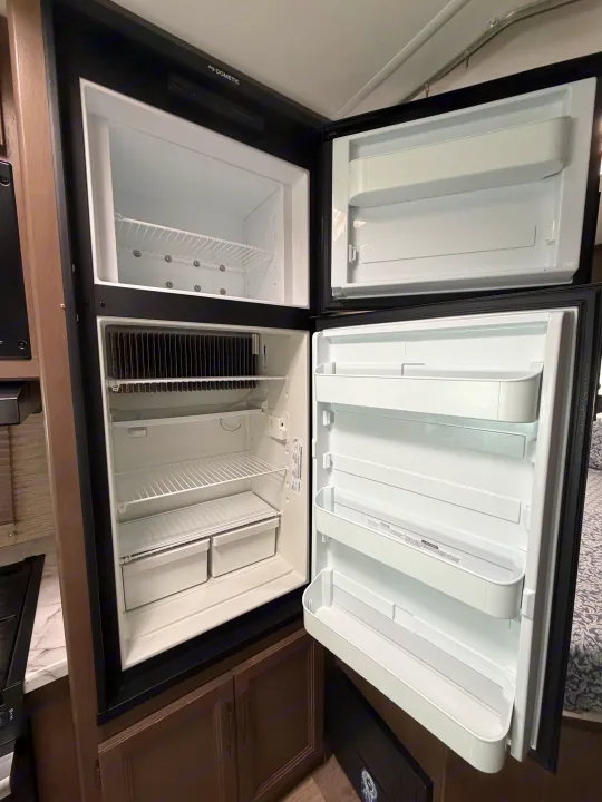 Clean Dometic RV rental refrigerator interior, doors open, showing an empty freezer and main compartment with shelves and drawers.. Thor Coleman 2023