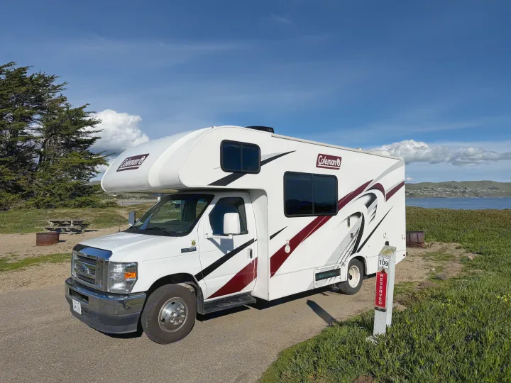 A white Coleman RV rental is parked at reserved campsite 109, offering scenic views of the ocean and distant hills under a blue sky.. Thor Coleman 2023