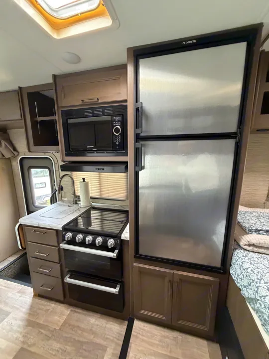 Inside an RV rental: Compact kitchen featuring a Dometic fridge, microwave, stove/oven, sink, and cabinetry, next to the bed.. Thor Coleman 2023