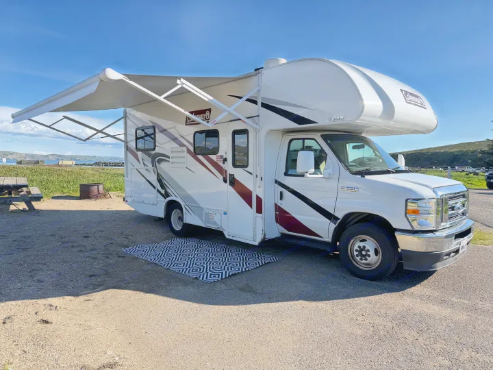 An RV rental, a white motorhome with an extended awning and geometric rug, parked at a scenic coastal campsite.. Thor Coleman 2023