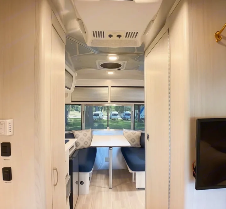 Step inside this modern RV rental's bright living space, featuring a spacious dinette with blue cushions and large windows overlooking other RVs.. Airstream Bambi 2024