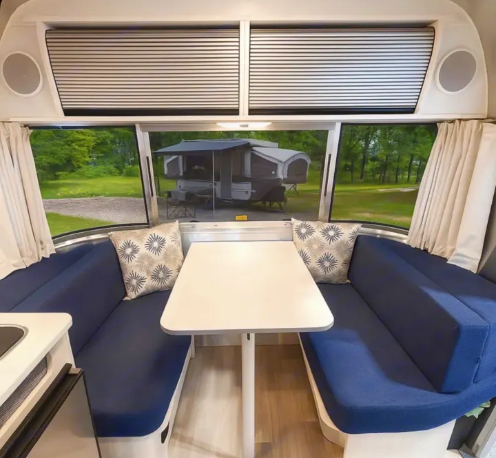 Inside a bright rv rental, a dining area with blue bench seats, a white table, and a scenic view of a campsite and another camper.. Airstream Bambi 2024