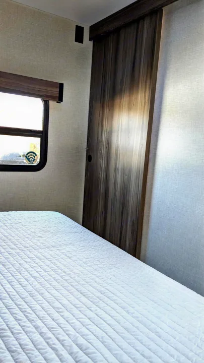 Cozy RV rental bedroom featuring a white quilted bed, window, and a modern wooden sliding door, offering a comfortable interior.. KZ Connect SE 2025