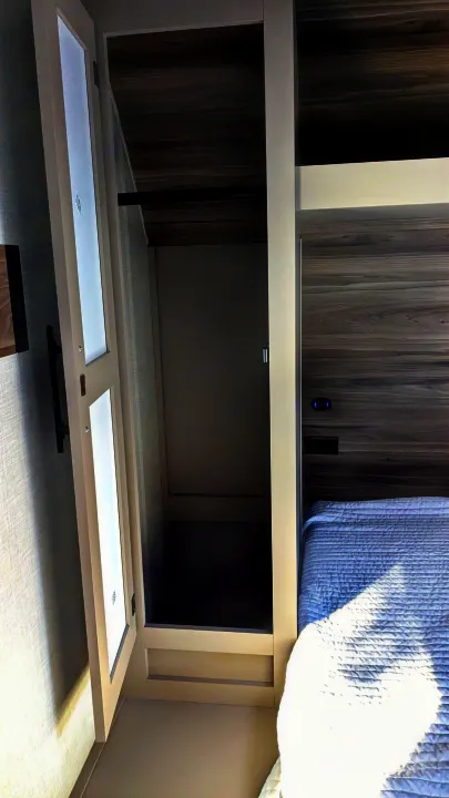 RV rental interior: an open closet with a hanging rod, adjacent to a bed with a white quilt and dark wood-paneled wall.. KZ Connect SE 2025