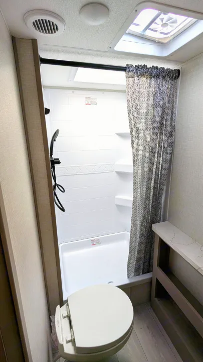 RV rental bathroom features a white shower with patterned curtain, compact toilet, and practical side shelving for convenience.. KZ Connect SE 2025