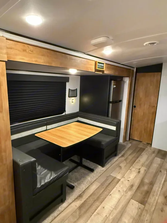 Modern RV rental dinette with black booth seating, wooden table, dark window blinds, and a built-in refrigerator.. Keystone RV Springdale 2022