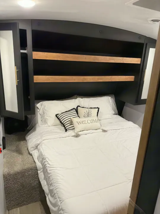 Cozy RV rental bedroom featuring a white bed, modern black overhead cabinets with wood shelves, and ample storage for comfortable travel.. Keystone RV Springdale 2022