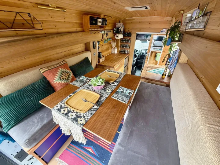 Cozy RV rental's wooden interior, featuring a dining table set for two, cushions, a kitchenette, and forward view.. Ford Transit 2019