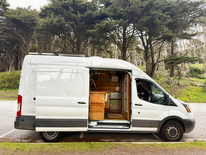 Experience van life! This white rv rental campervan, open to show its wooden interior and kitchen, is parked by lush cypress trees.. Ford Transit 2019