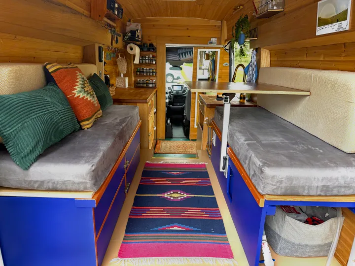 Cozy RV rental interior: wood-lined, vibrant blue storage, grey cushions, colorful pillows, and a compact kitchen. Road trip ready!. Ford Transit 2019