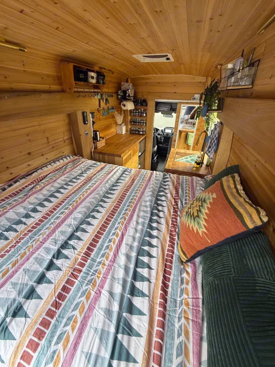 Cozy RV rental interior with a patterned bed, warm wood paneling, and a compact kitchen area with sink and storage.. Ford Transit 2019