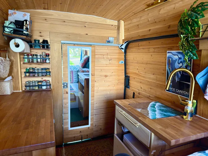 Inside this rv rental, a compact wooden kitchen with a sink, organized spice racks, and shelves create a cozy van interior.. Ford Transit 2019