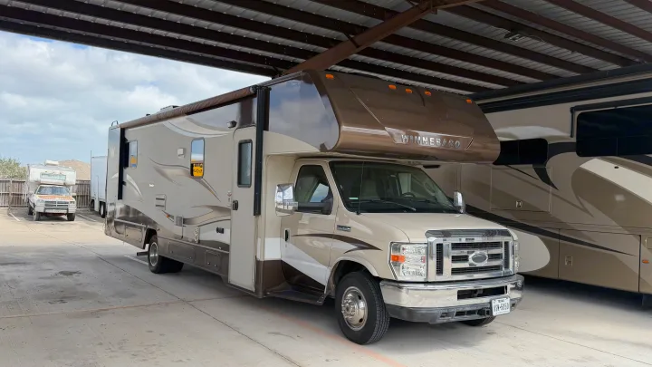 Beige & brown Winnebago Class C motorhome for RV rental, parked under a canopy with other RVs. Perfect for your next adventure.. Winnebago Minnie Winnie 2015