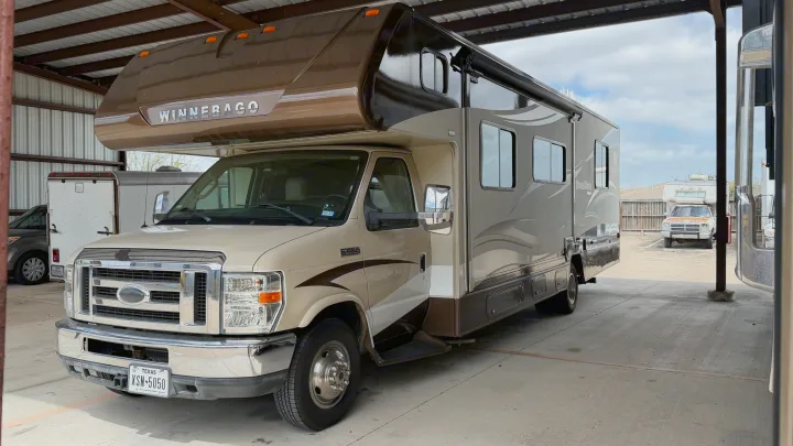 Cream & brown Winnebago Minnie Winnie RV rental, parked under a shelter. Front-side view with Texas plate XSN-5050.. Winnebago Minnie Winnie 2015