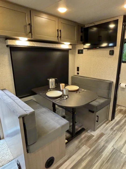 Bright RV rental dining area: a table with plates, cups, and kettle between two comfy upholstered benches. Overhead cabinets & TV.. Jayco Jay Feather Air 2025