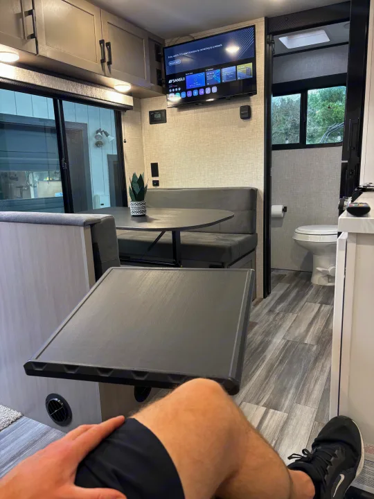 Modern RV rental interior showing a person's legs, a dinette with a plant, smart webOS TV, and a visible bathroom.. Jayco Jay Feather Air 2025