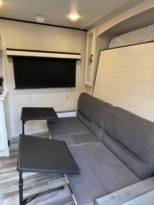 Inside an RV rental: a cozy gray sofa, adjustable tables, a window blind, and a space-saving murphy bed.. Jayco Jay Feather Air 2025