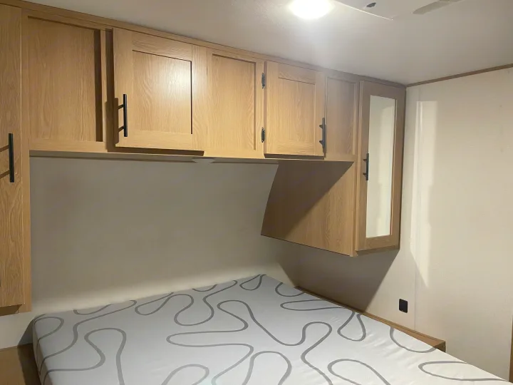 Inside an RV rental bedroom: light wood overhead cabinets, one with a mirror, above a bed with a grey patterned mattress.. Avenger Avenger Trailer 2022