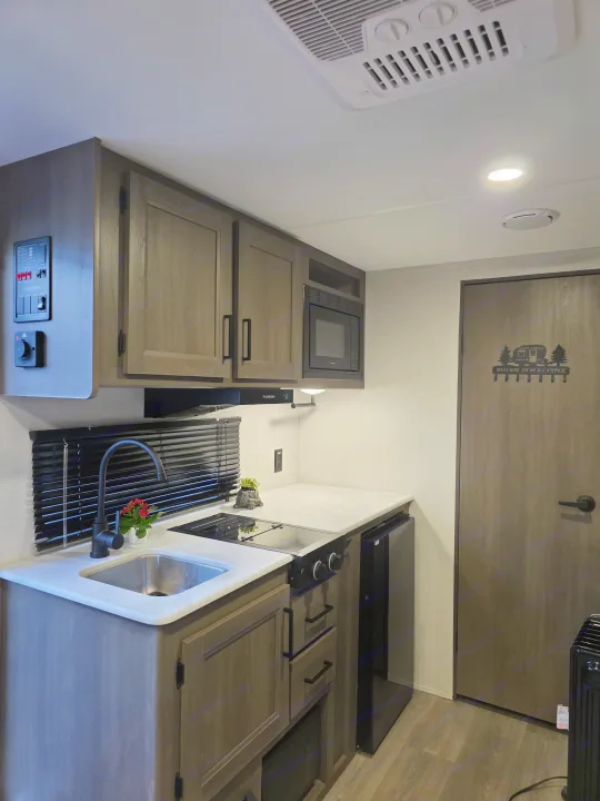 This modern RV rental kitchen features a sink, stovetop, microwave, and fridge, all within a well-appointed camper interior.. Grand Design Transcend One 2025