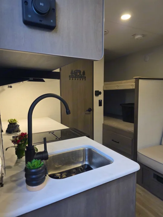 Modern RV rental kitchen with a black faucet, sink, and cooktop. Bunk beds are visible in the background for a complete camper experience.. Grand Design Transcend One 2025